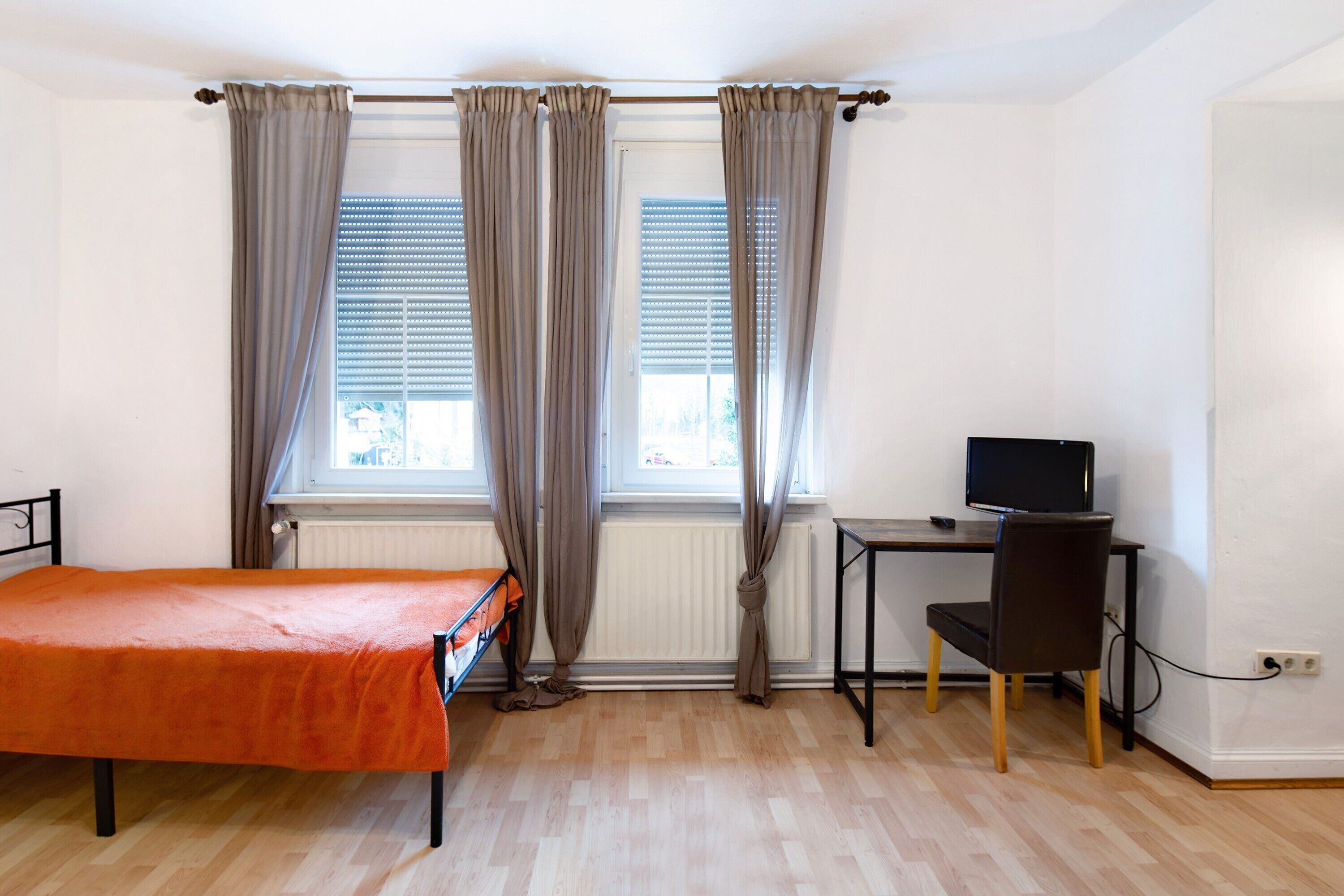 A room with a bed, a desk with a chair, a television, and two windows with curtains.
