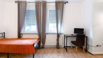 A room with a bed, a desk with a chair, a television, and two windows with curtains.