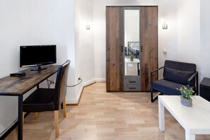 Ein Zimmer mit Schreibtisch aus Holz, einem Sessel, einer Couch, einem kleinen Tisch mit Pflanze und einem Kleiderschrank mit Spiegel.