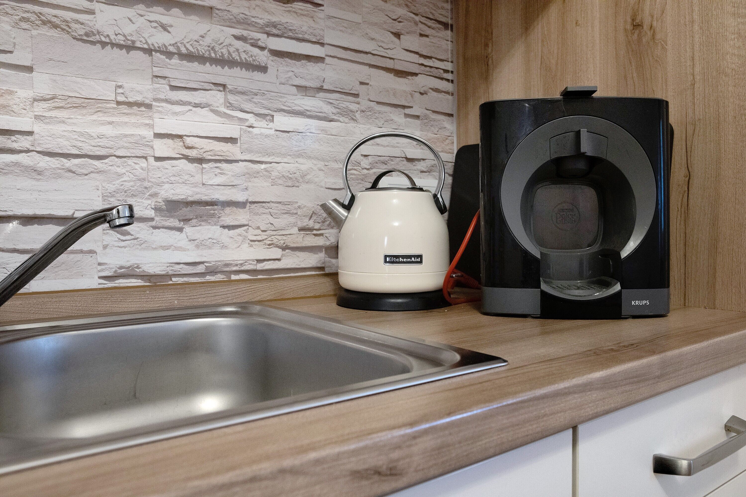A kitchen with a modern design, featuring a stone-textured backsplash, a stainless steel sink, a white kettle, and a Krups coffee maker.
