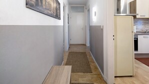 Un couloir étroit avec un banc en bois, une porte au fond et un coin cuisine avec des placards et un réfrigérateur.