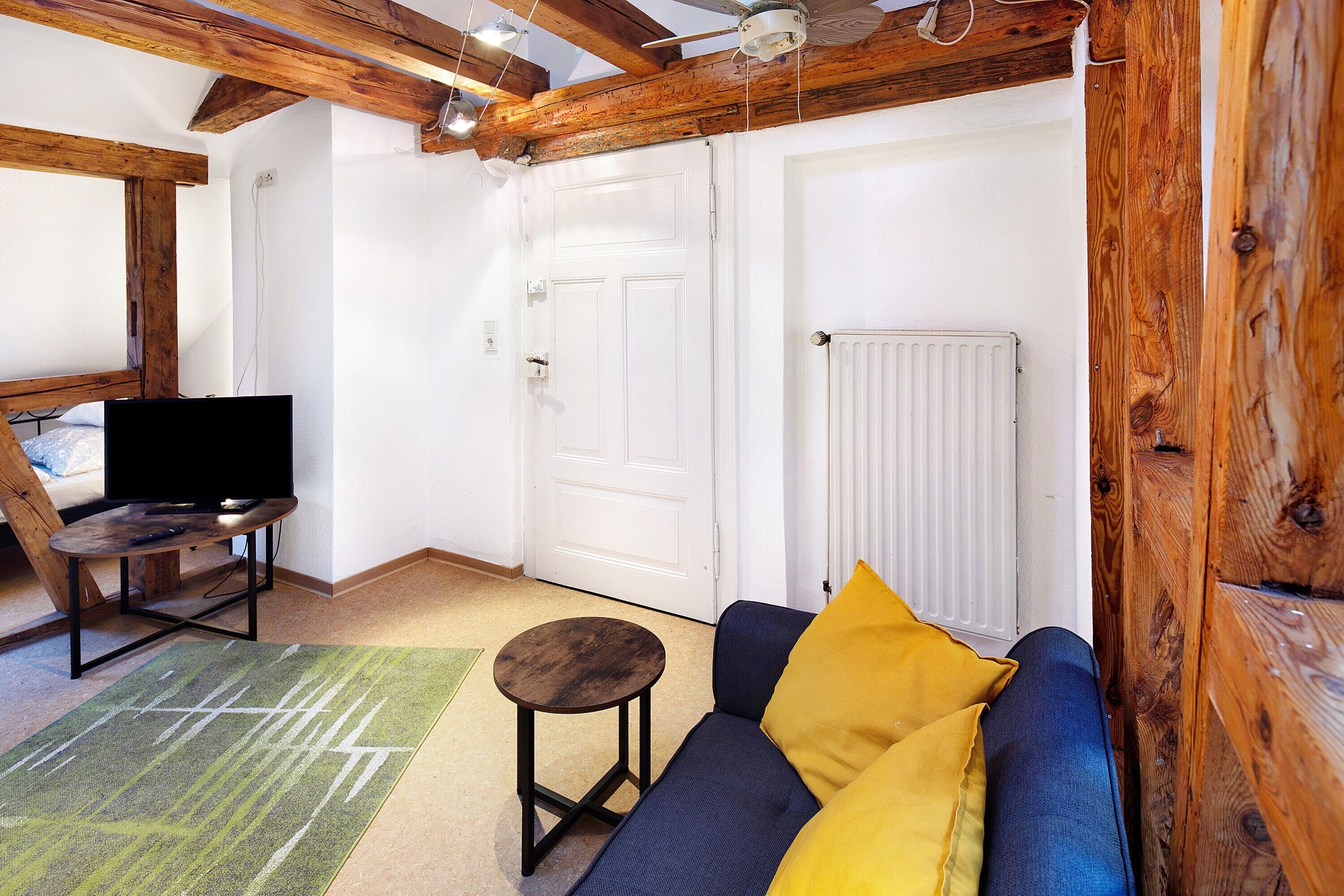 A room with a sofa, a television on a stand, a door, and wooden beams on the ceiling.