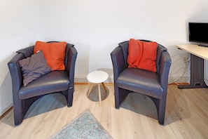 Deux fauteuils en cuir avec des coussins rouges, une petite table blanche et un téléviseur posé sur un bureau, dans une pièce dont le sol est recouvert de parquet.