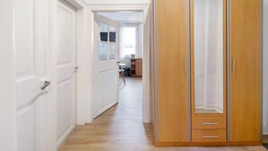 Un couloir avec des portes blanches, une armoire en bois dotée de deux tiroirs, et une vue sur une pièce avec une fenêtre et un bureau.