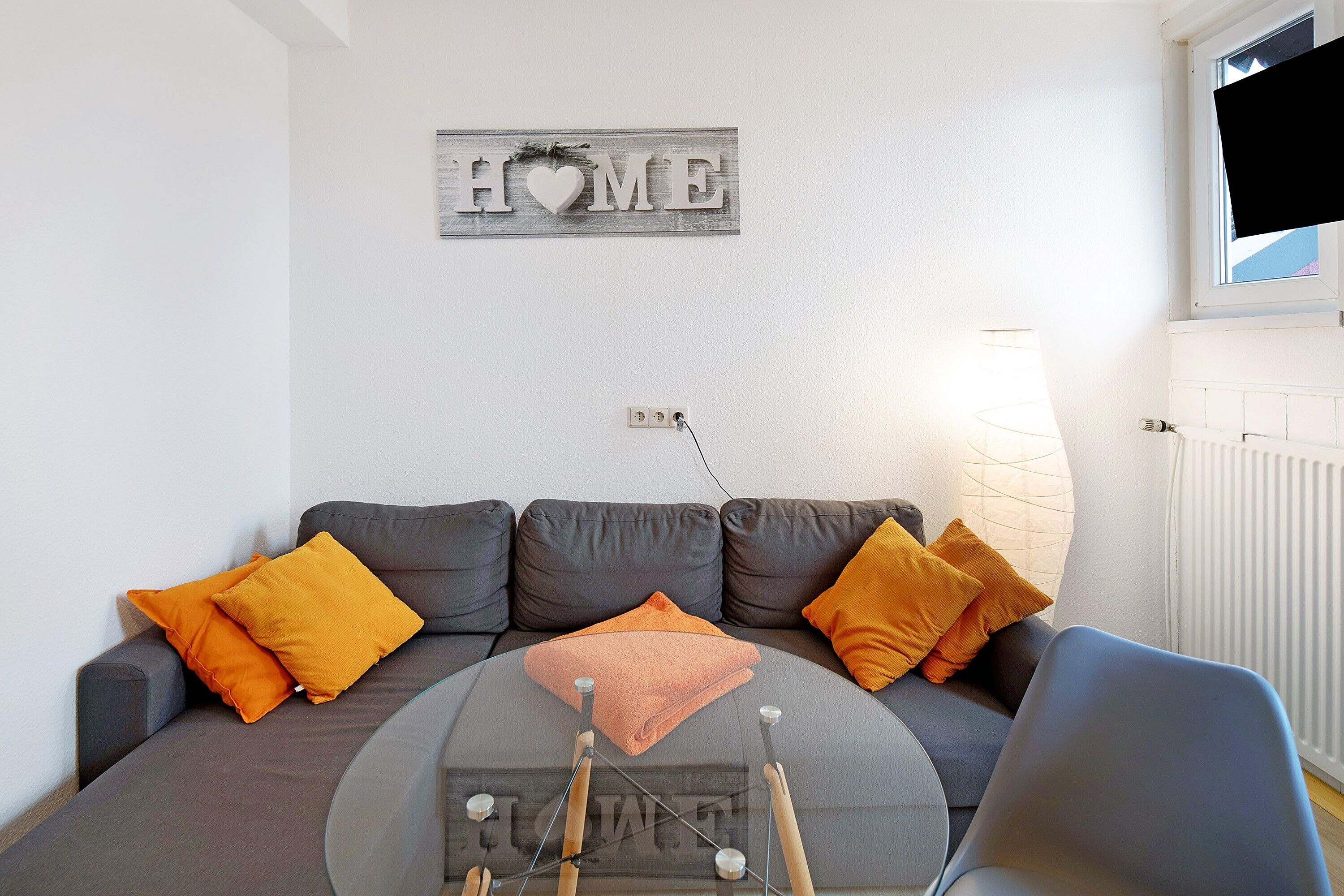 Olohuone, jossa on harmaa sohva, oranssit koristetyynyt, pyöreä sohvapöytä ja seinällä oleva koristeellinen kyltti, jossa lukee ”HOME”.