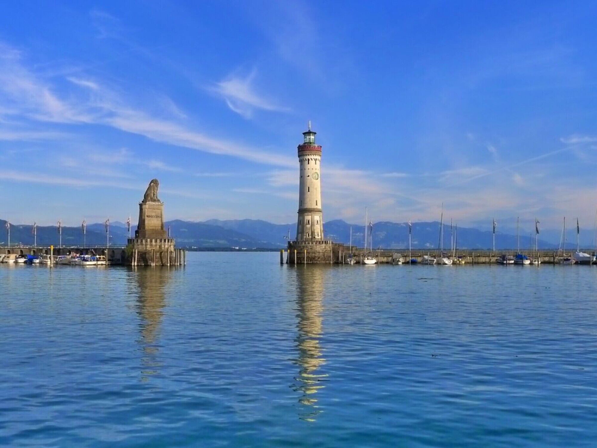 Een vuurtoren en een standbeeld in een rustige haven, met boten en bergen op de achtergrond.