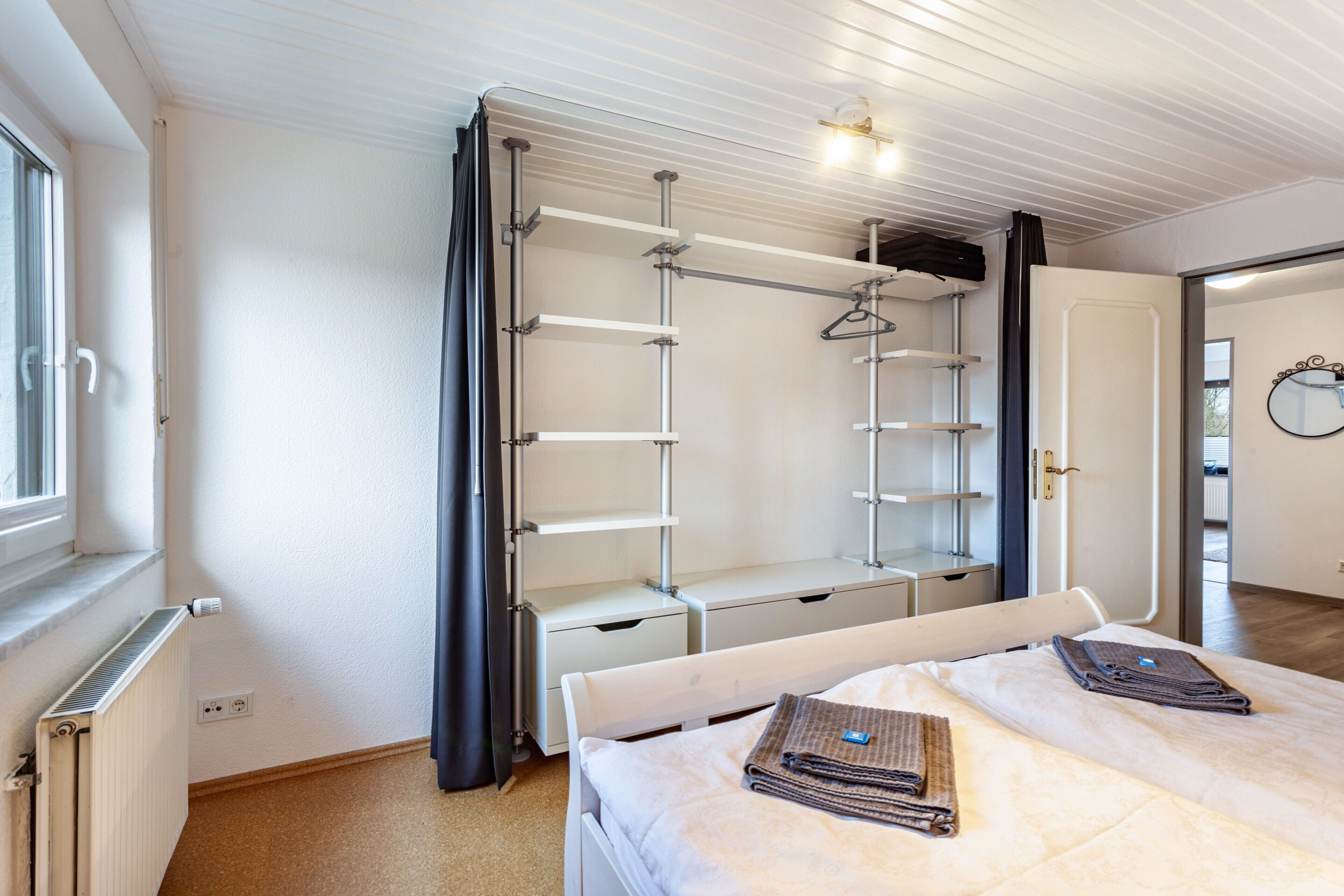 Een slaapkamer met een bed, een radiator, een kledingkast met planken en een deur naar een andere kamer.