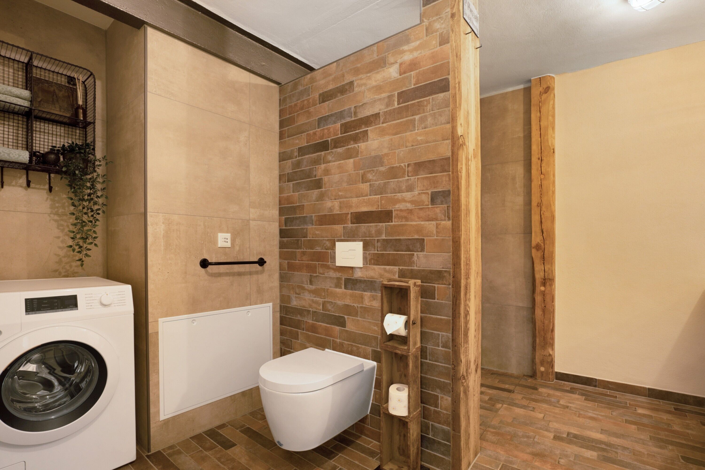 Une petite salle de bain équipée d’un lave-linge, de toilettes et d’une étagère en bois où sont rangés les articles de toilette.