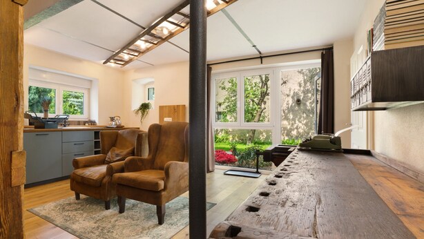 Una cocina moderna con encimera de madera, un sillón de cuero marrón y vistas a un jardín a través de amplios ventanales.