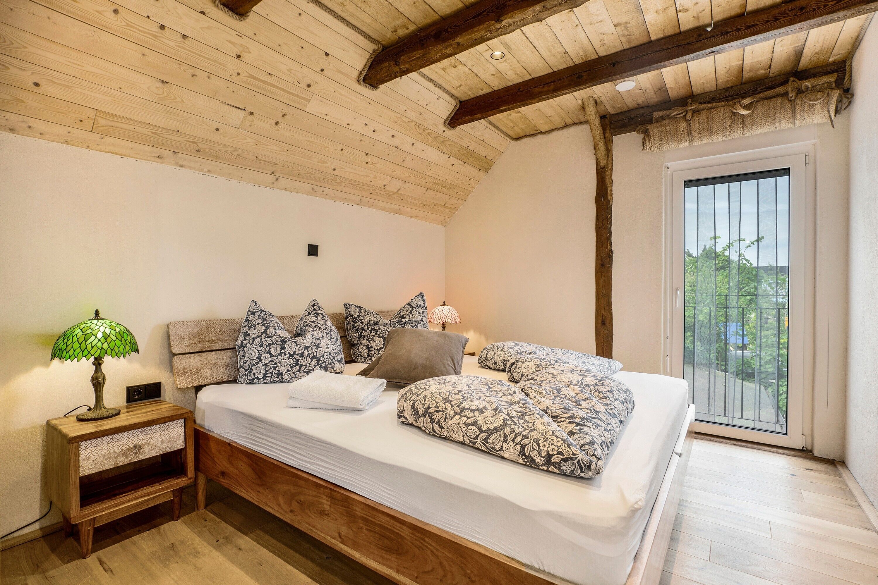 Un dormitorio con techo de madera, cama con ropa de cama estampada, mesita de noche con lámpara y vista por la ventana.