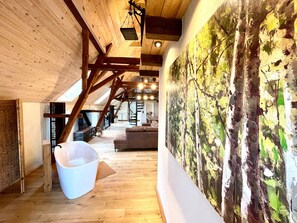 Un intérieur avec des poutres apparentes, un seau blanc, un tableau représentant des arbres et un escalier.