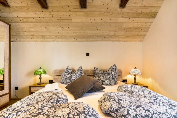 Une chambre avec un plafond en bois, un lit recouvert d’une literie à motifs floraux, deux tables de chevet avec des lampes et un miroir sur le mur de gauche.