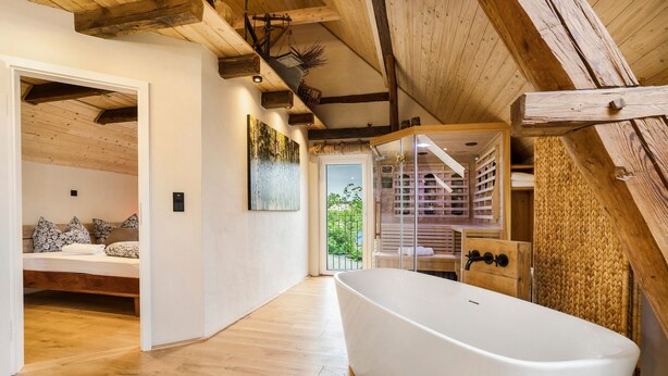 Ein modernes Interieur mit Holzboden, einer weißen Badewanne, einem Bett mit gemusterten Kissen und Blick auf einen Garten durch eine Glastür.