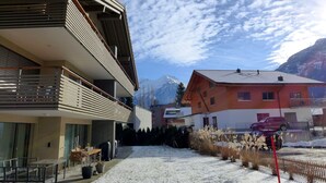 Un edificio moderno con un balcone, una zona salotto esterna coperta e vista sulle montagne innevate.