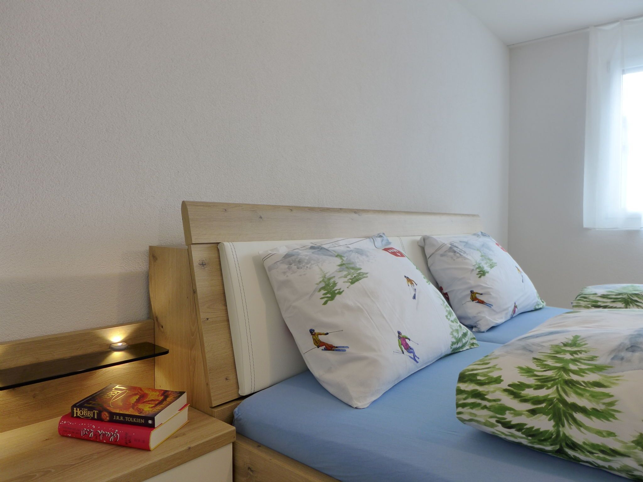 A bedroom with a wooden headboard, a bed with blue bedding and patterned pillows, a bedside table with books, and a window with white curtains.