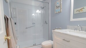 Une salle de bain dotée d’une douche à parois de verre, de toilettes blanches, d’un meuble-lavabo avec un évier et deux robinets, et d’un miroir au-dessus du meuble-lavabo.