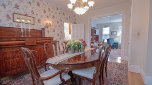 Uma sala de jantar com mesa de madeira, cadeiras e papel de parede floral. Há um candelabro sobre a mesa e um tapete estampado no chão.