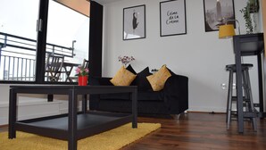 A modern living room with a black sofa, a coffee table, and a yellow rug. There are framed pictures on the wall and a plant on a shelf. - Penthouse Sleeps 2 with Stunning Views (Birmingham)