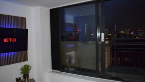 A modern living room with a large window, a flat-screen TV mounted on the wall, and a small table with a plant. - Penthouse Sleeps 2 with Stunning Views (Birmingham)