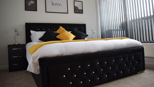 A bedroom with a black upholstered headboard, white and yellow bedding, a black nightstand with drawers, and a window with vertical blinds. - Penthouse Sleeps 2 with Stunning Views (Birmingham)