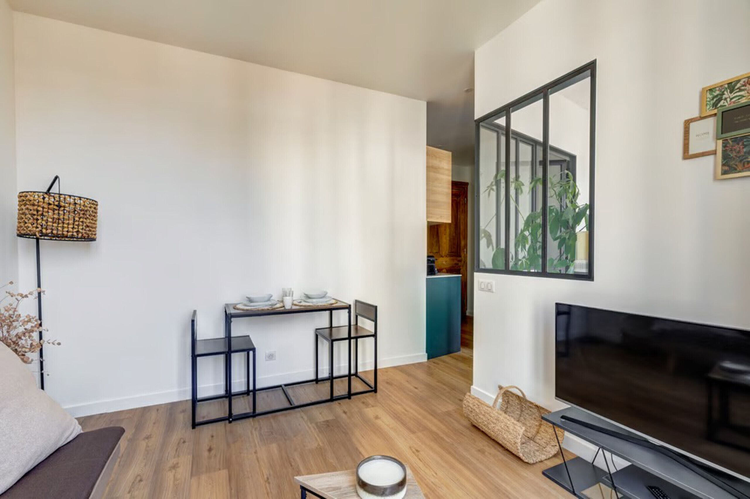 A modern living room with a flat-screen TV, a dining area with a table and chairs, and a kitchen visible through an open doorway.