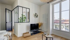 Un salon moderne doté d’un téléviseur à écran plat, d’un coin repas avec une table dressée pour quatre personnes et d’une grande fenêtre offrant une vue sur la ville.