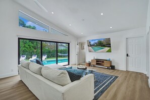 A modern living room with a large sofa, a TV mounted on the wall, and a view of a swimming pool.