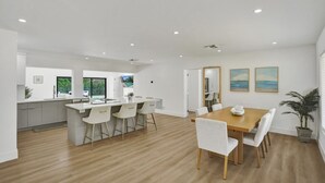 Una cocina moderna con una isla central, un comedor con mesa de madera y sillas blancas, y vistas al exterior a través de un amplio ventanal.