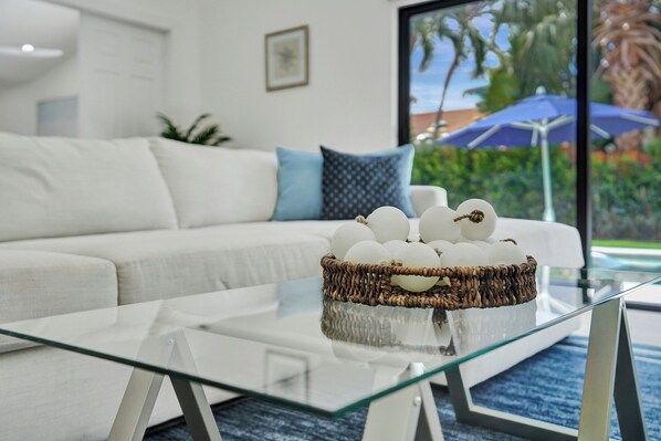 A modern living room with a glass coffee table, a white sofa with blue cushions, and a wicker basket with decorative items.