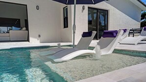 Una piscina con sillas de descanso y una sombrilla, un edificio blanco con puerta de cristal y vistas al exterior.