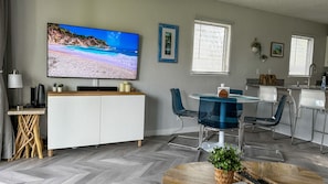 A modern living room with a flat-screen TV, a dining area with blue chairs, a kitchen with white cabinets, and a herringbone-patterned floor.