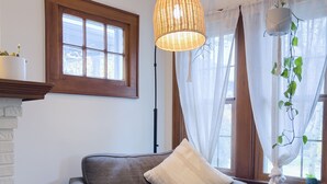 Una sala de estar acogedora con un sofá gris, una mesita de mimbre, una lámpara de pie y una ventana con cortinas translúcidas.