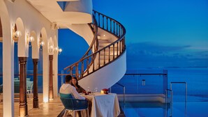 Um casal jantando à mesa com vista para o mar, uma escadaria em espiral e uma piscina.