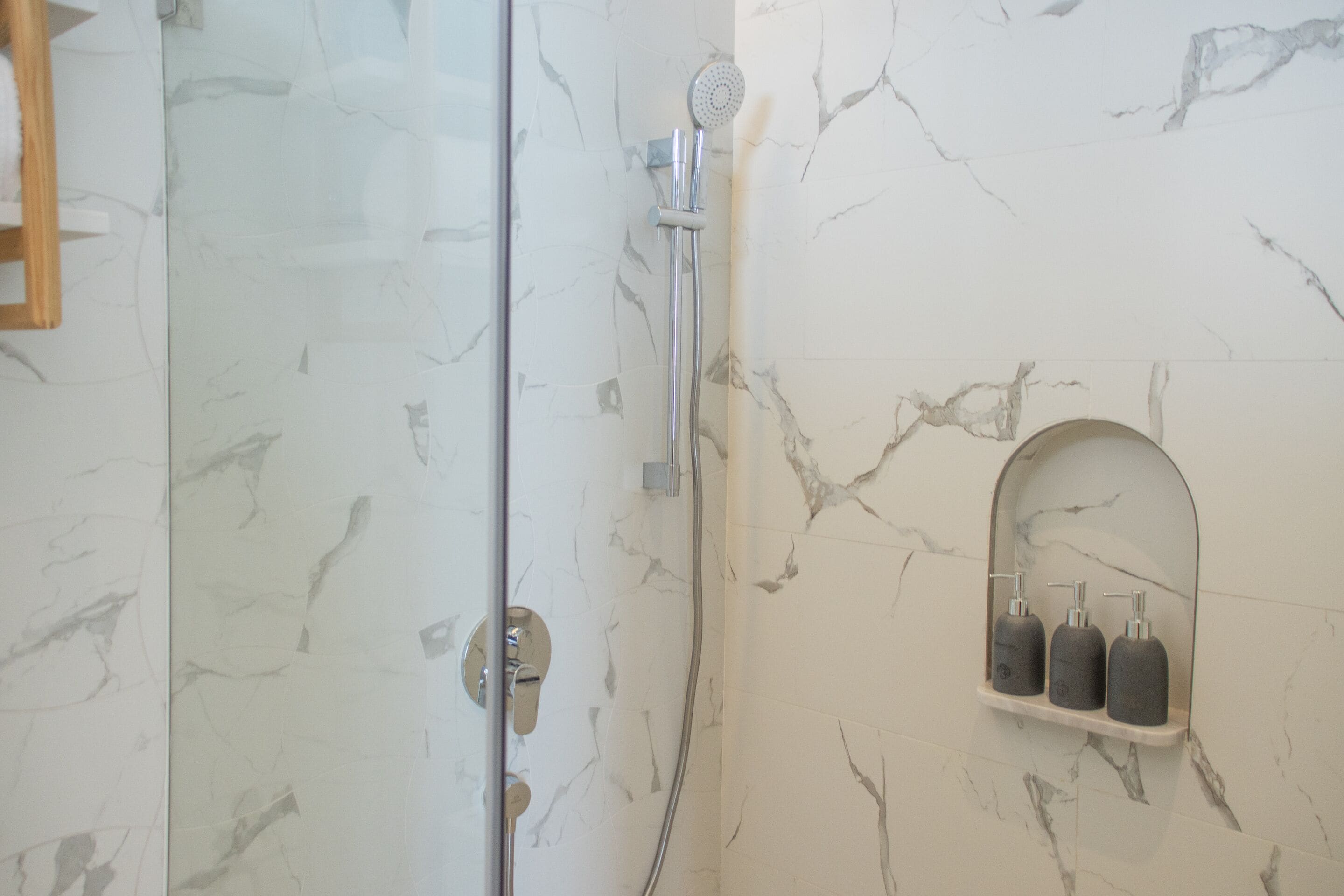 A bathroom with a glass shower door, a handheld showerhead, and a marble wall with a built-in soap dispenser shelf.