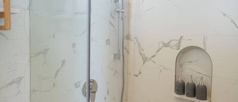 A bathroom with a glass shower door, a handheld showerhead, and a marble wall with a built-in soap dispenser shelf.