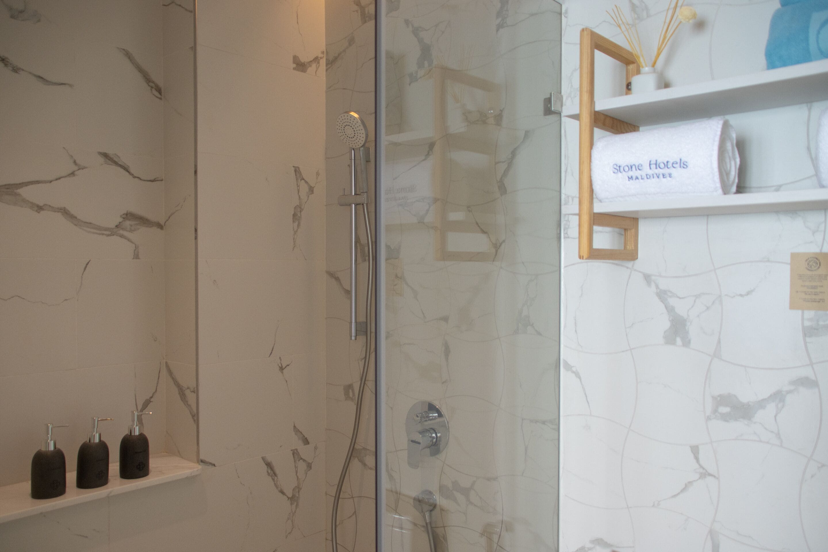 Un baño con cabina de ducha de vidrio, paredes de mármol y un estante con artículos de aseo y una toalla que dice 'Stone Hotels Maldives'.