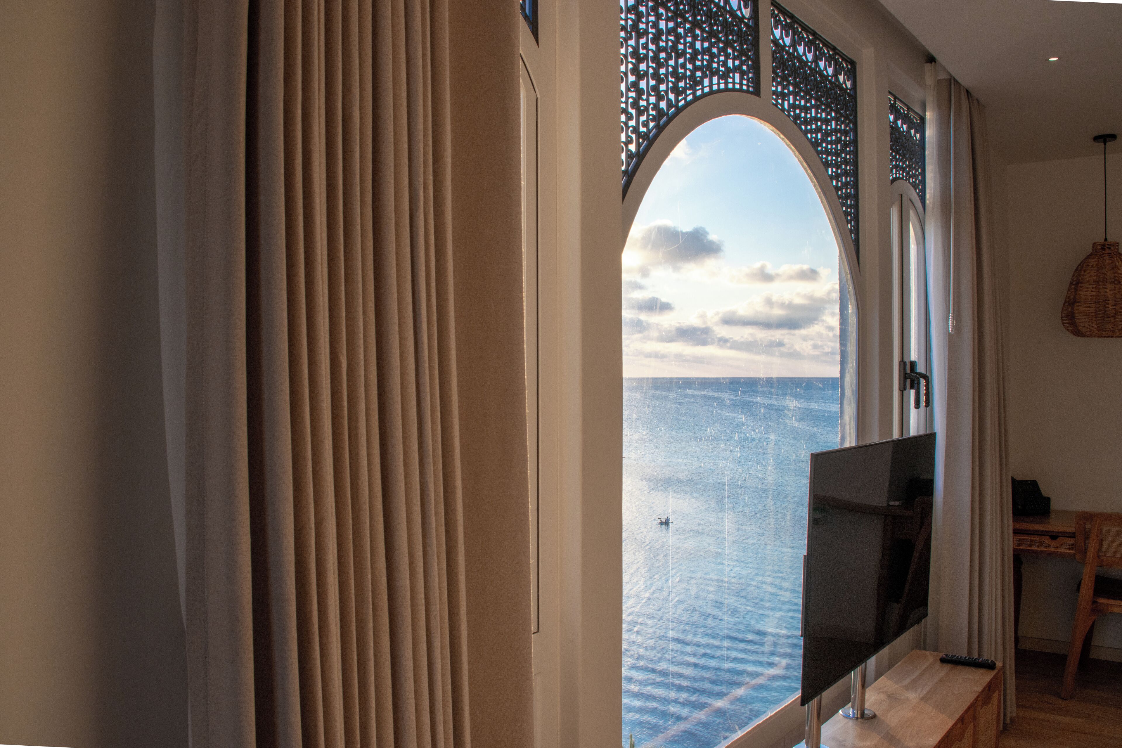 A room with a large arched window overlooking the sea, a flat-screen TV, and a wooden desk.
