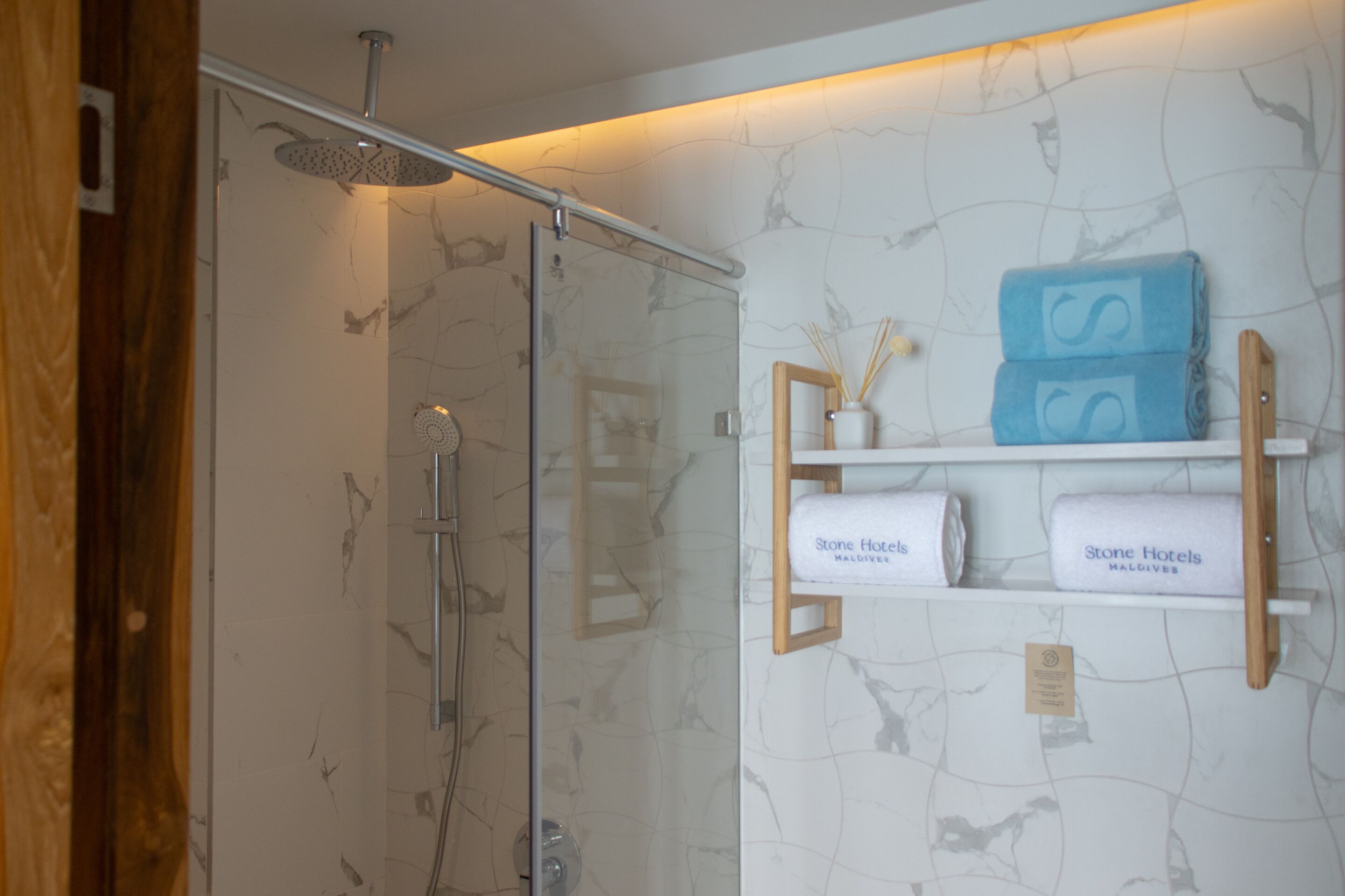 Un baño con cabina de ducha de vidrio, paredes de mármol y estantes con toallas que dicen "Stone Hotels Maldives".