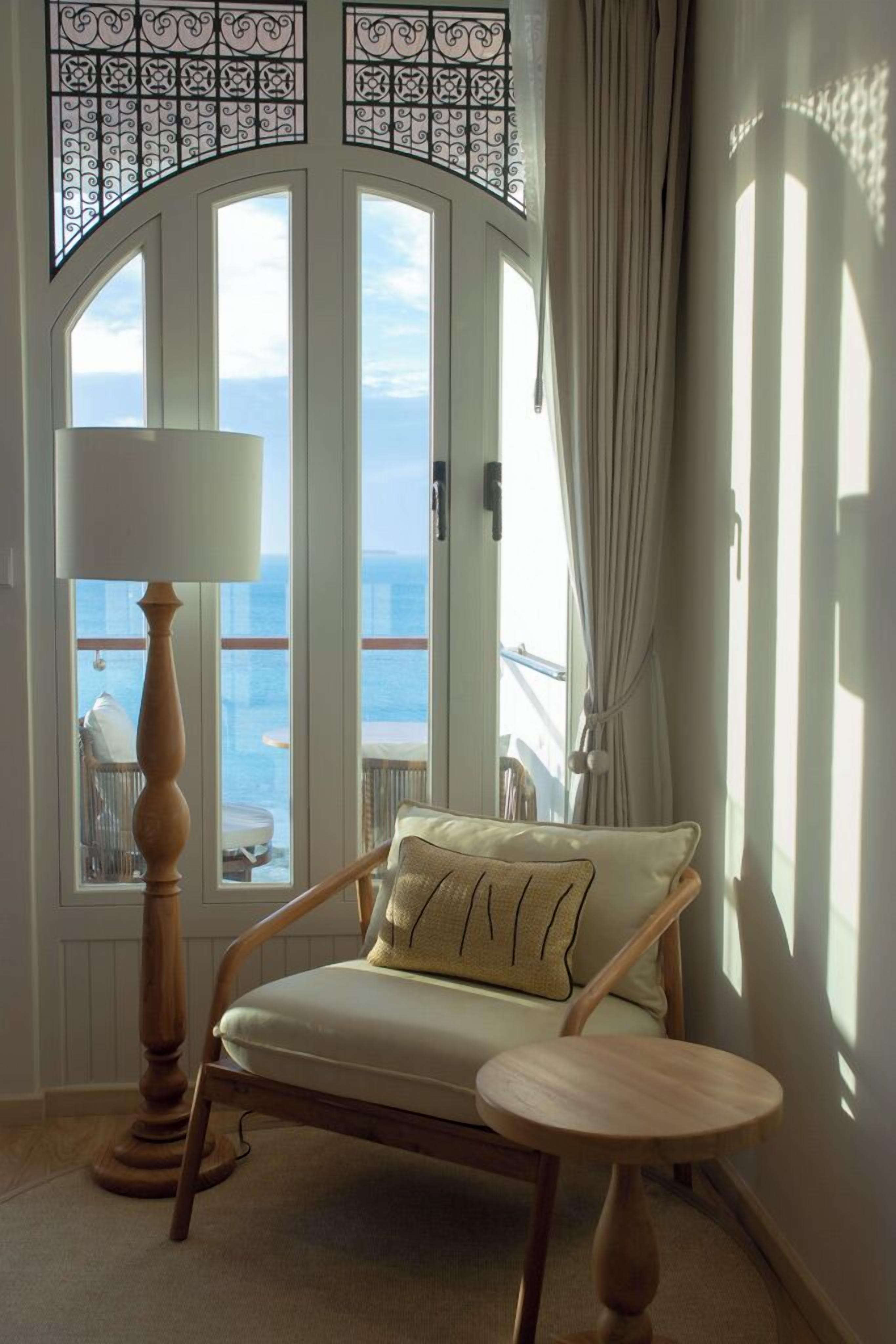 Una habitación con vista al mar, una silla de madera con cojín, una mesita redonda y una lámpara de pie.