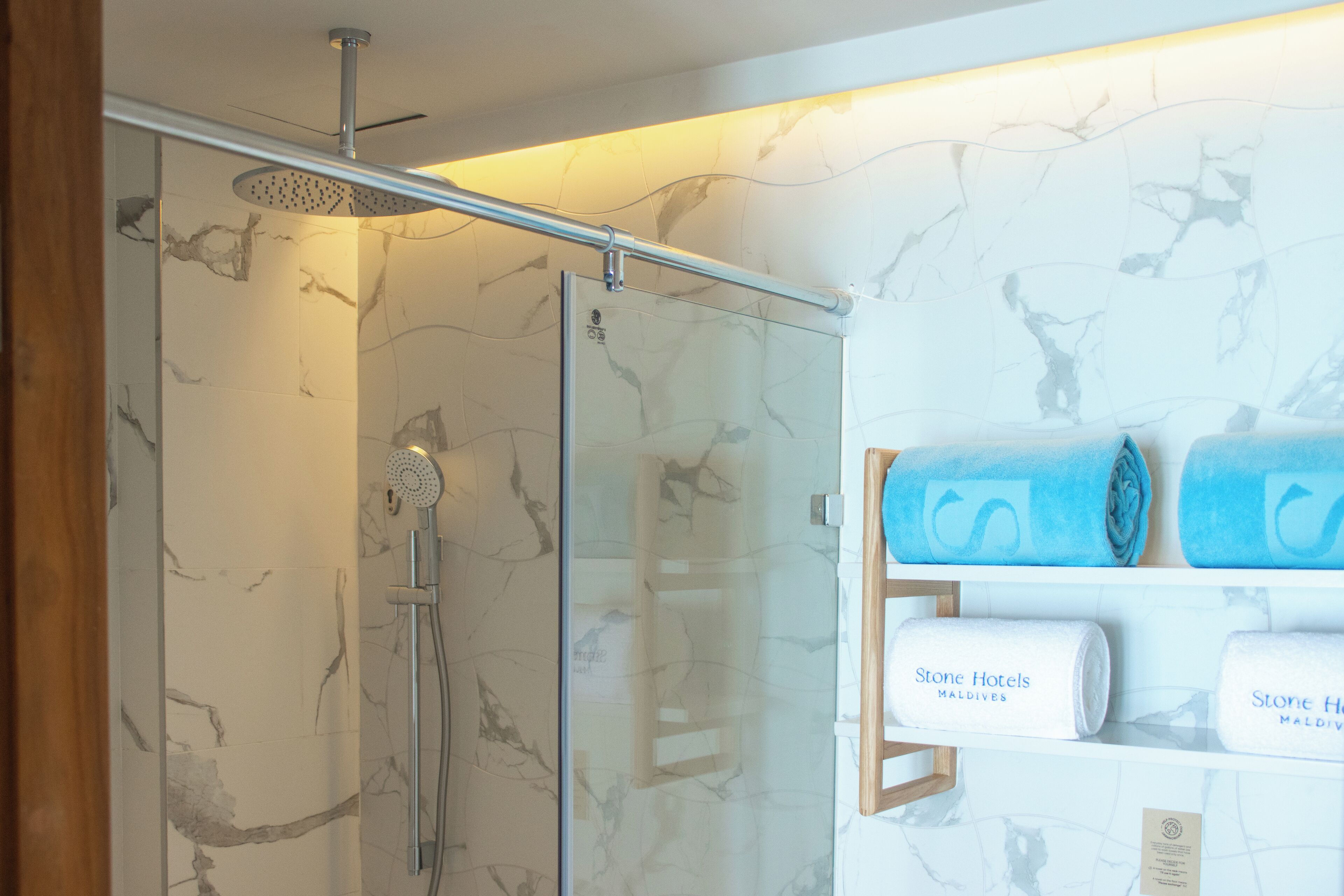 A glass-enclosed shower with a handheld showerhead, a glass door, and a marble-patterned wall. There are towels with "Stone Hotels Maldives" branding on a shelf.
