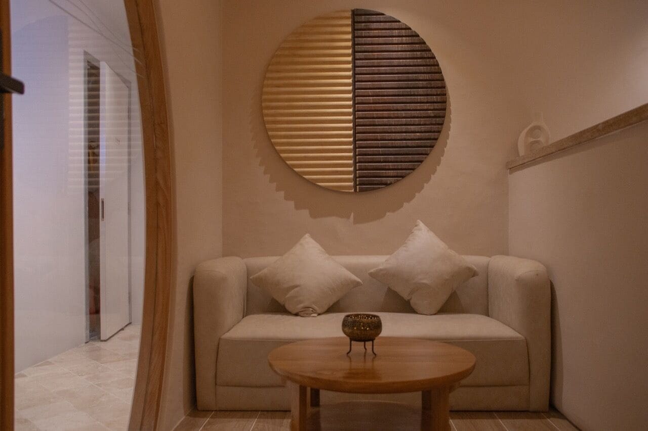 Un soggiorno moderno con un divano beige, due cuscini bianchi, un elemento decorativo rotondo a parete e un piccolo tavolino in legno.