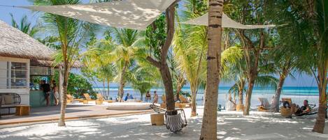 Un complejo turístico frente a la playa con cabañas de techo de paja, palmeras y sillones de sol.