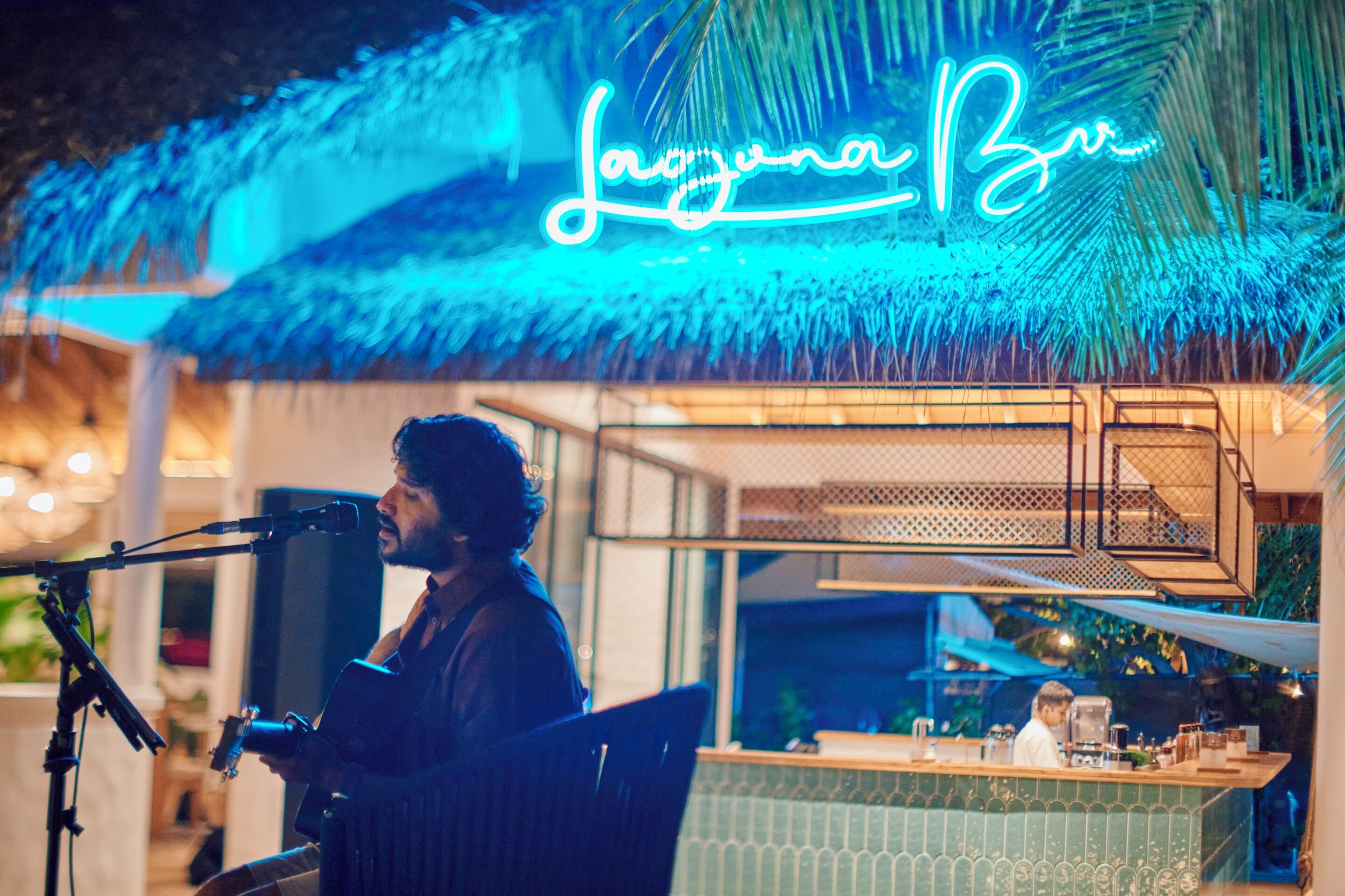 Une personne joue de la guitare dans un bar, et une enseigne au néon indique « Laguna Bar ».