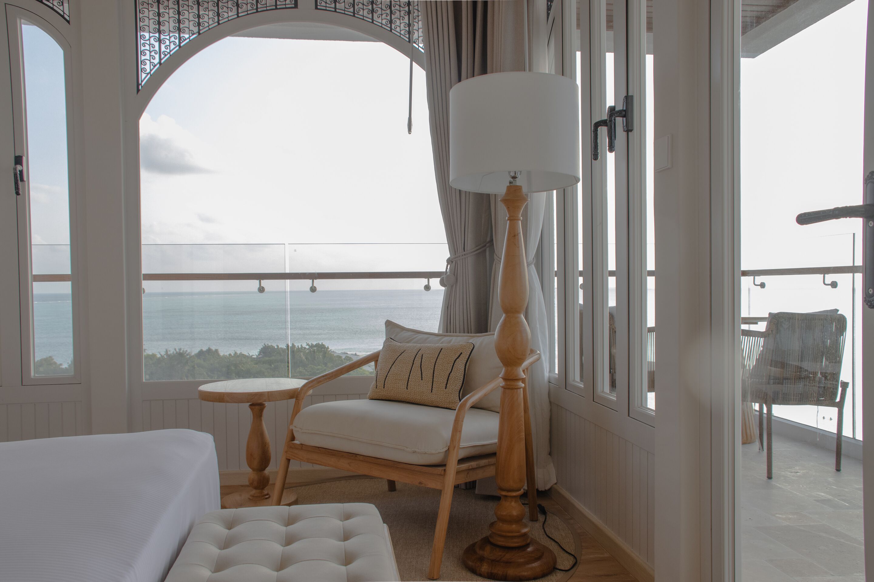 Una habitación con una cama, un sillón blanco con cojín, una mesita y una lámpara de pie. Cuenta con un balcón con vista al mar.