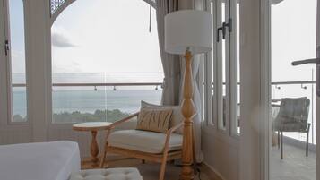 Una habitación con una cama, un sillón blanco con cojín, una mesita y una lámpara de pie. Cuenta con un balcón con vista al mar.