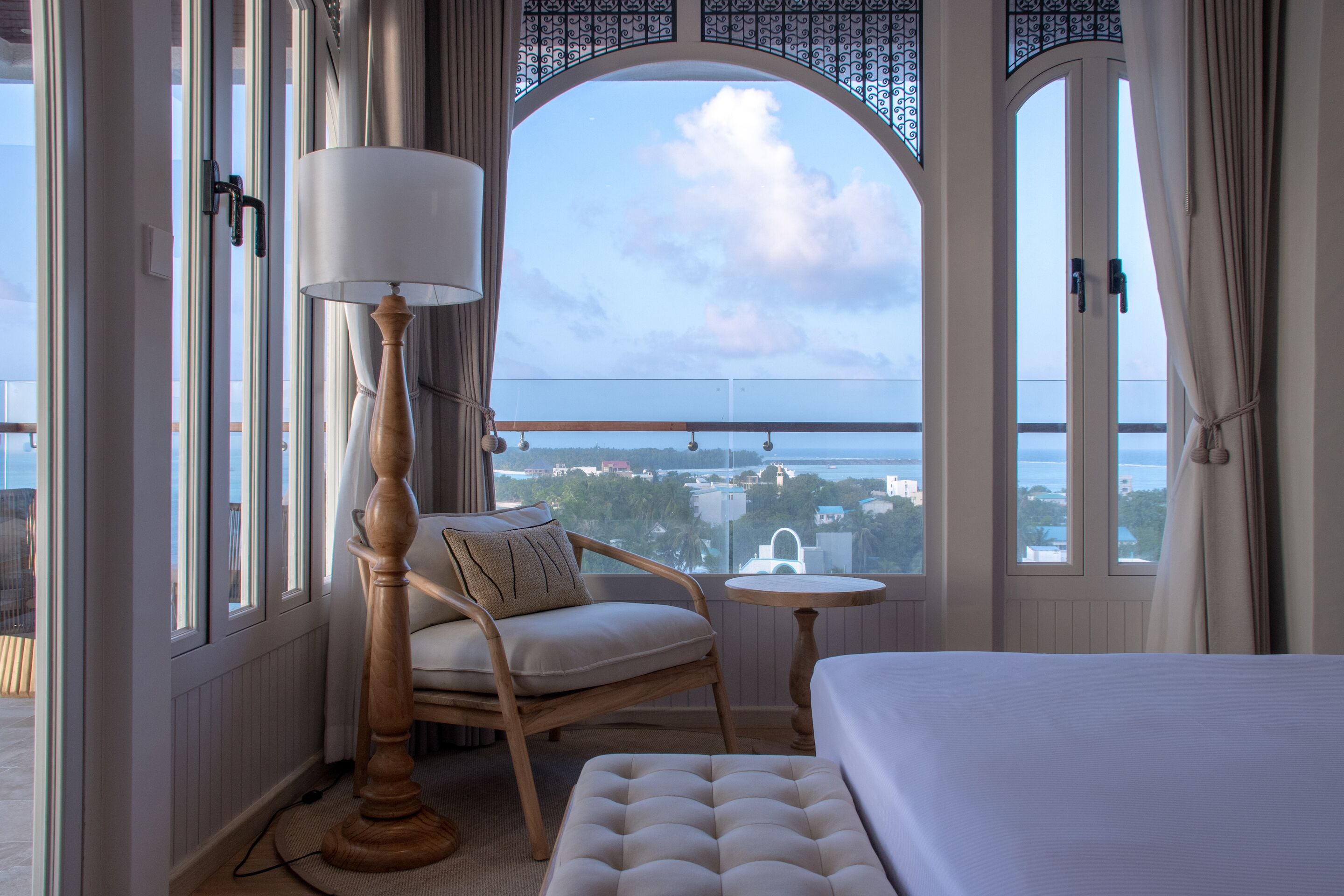 A hotel room with a view of the sea, a bed, a chair, a lamp, and a small table.