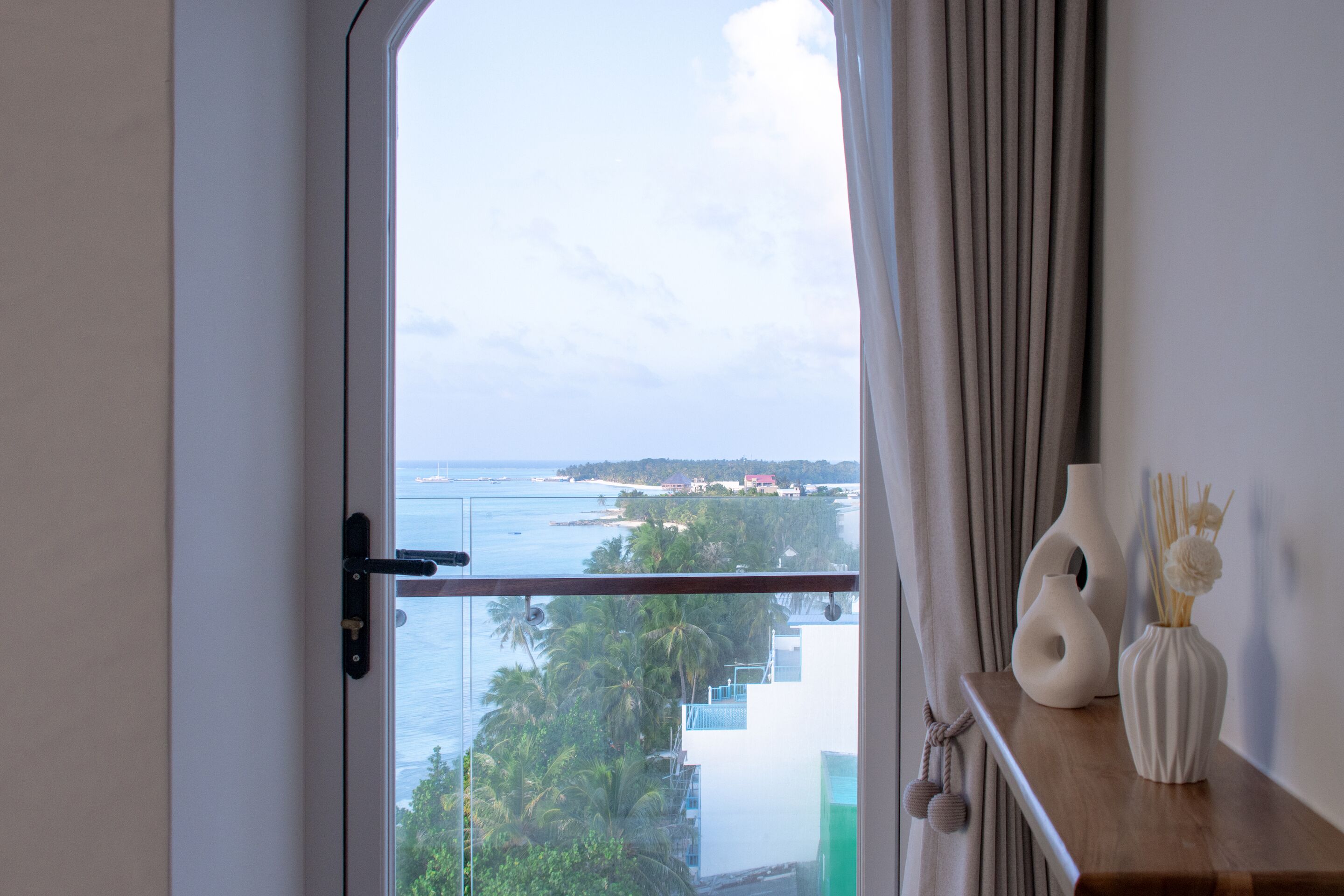 A room with a view of the ocean and palm trees, a wooden shelf with vases, and a curtain partially drawn.