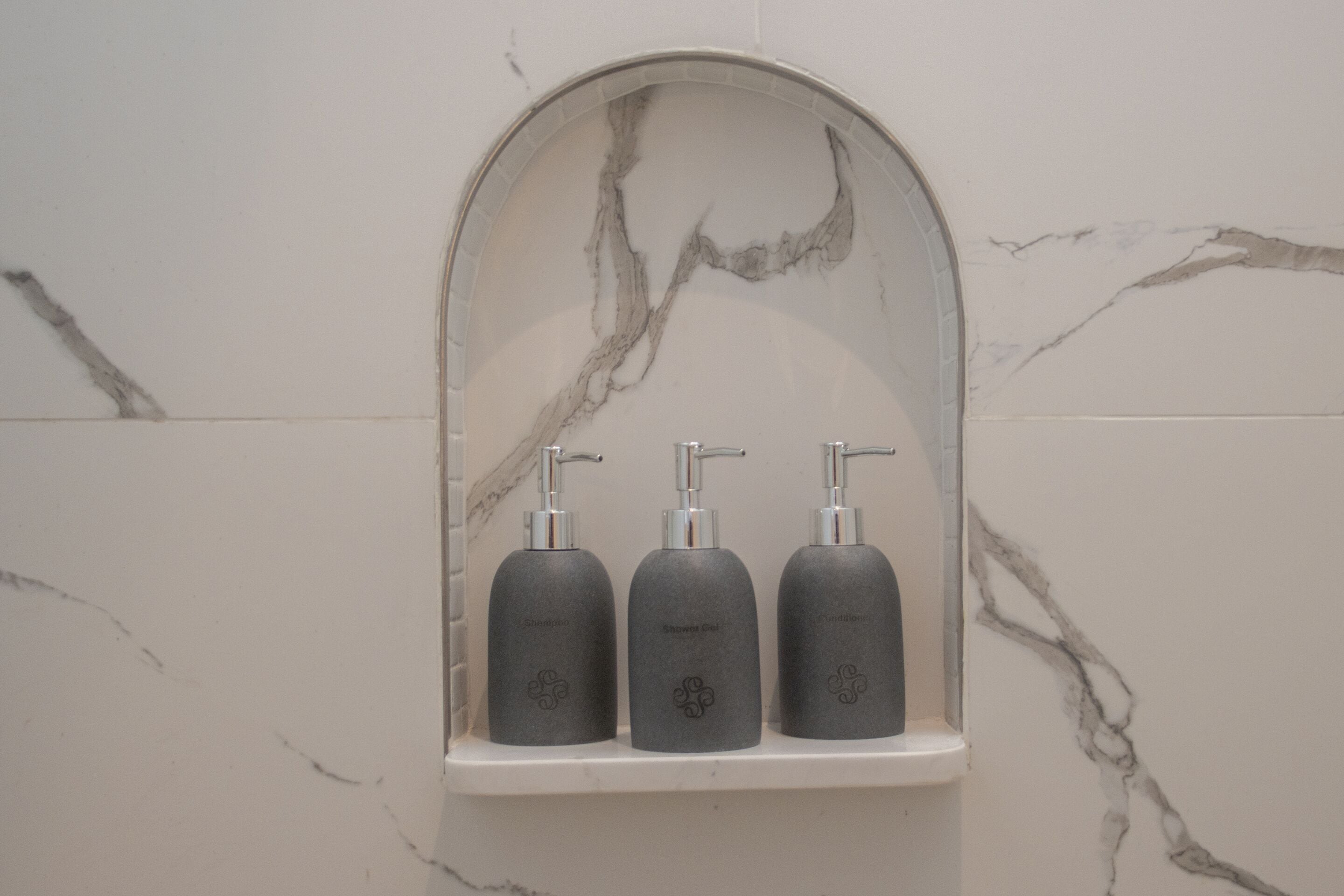 A bathroom with a marble wall and a shelf holding three bottles of shampoo, conditioner, and body wash.