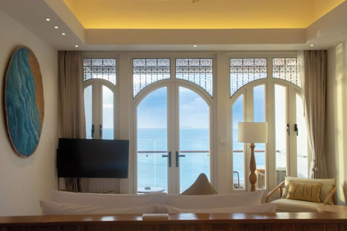 Hotellihuone, jossa on suuri, kaareva ikkuna, josta avautuu näkymä merelle, seinälle asennettu taulutelevisio, puinen yöpöytä lampun kanssa sekä mukava oleskelualue tyynyineen.