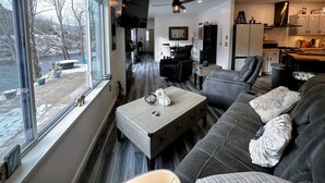 A modern living room with a large window, a sofa, a coffee table, and a kitchen area visible through an open door. - 37PA Superbly appointed riverfront home in LIttleton! Skiing, hiking, firepit, wifi, dog friendly! (Littleton)